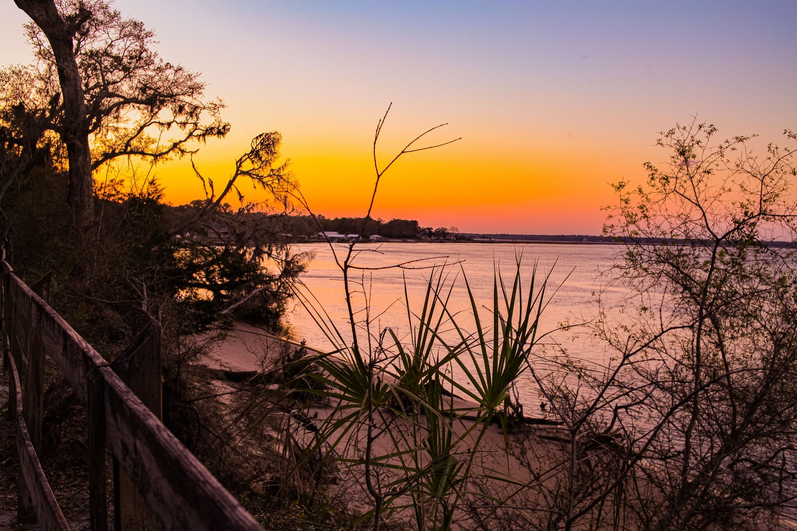 A serene water view at sunset with trees and a wooden fence in the foreground.